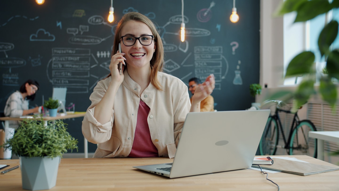 Mujer con gafas habla por teléfono en una oficina moderna.